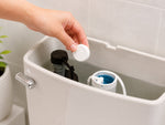 Hand placing a round white tablet into an open toilet tank next to flush mechanism, demonstrating product use