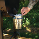 Hand placing a Pit Boss foil bucket liner into a small metal bucket attached to the side of a grill in an outdoor setting.