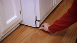 Close-up lifestyle photo of a hand placing a white tablet on a hardwood floor near a refrigerator base.
