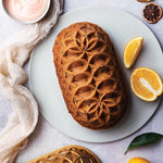 Decoratively patterned loaf cake served on a round plate with orange wedges, styling cloth, and the gold Jubilee loaf pan partially visible.