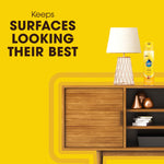 Pledge aerosol can next to a wooden sideboard and lamp with text ‘Keeps surfaces looking their best’ on yellow background.
