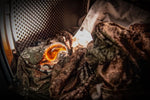 Laundry pod resting among camo clothing inside a dryer drum, lit dramatically to emphasize the orange gel capsule