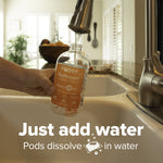 Hand holding a clear Podsy Bathroom cleaner bottle under a faucet being filled with water to dissolve a refill pod.