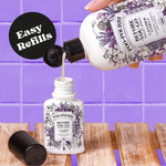Hand pouring liquid from a large Poo-Pourri lavender vanilla refill into a smaller Poo-Pourri bottle against a purple tiled background