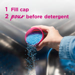 Hand pouring blue laundry scent crystals from a pink cap into a washing machine drum