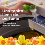 Hand pouring liquid fabric softener from a green cap into a washing machine dispenser with flowers along the bottom