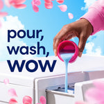 A hand pouring blue liquid from a pink cap into a washing machine dispenser with 'pour, wash, wow' text.