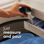 Hand pouring white liquid fabric softener from a measuring cap into a washing machine dispenser drawer.