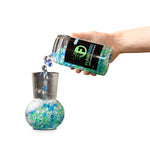 Hand pouring blue and green FunkAway beads from a clear jar into a ribbed glass vase to demonstrate use.