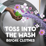 Hand pouring purple scent booster beads from a green measuring cup into a washer drum with floral illustrations and toss instruction text