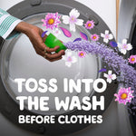 Hand pouring purple scent beads from a green measuring cap into a washing machine drum with floral overlay graphics