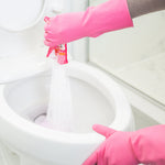 Gloved hands pouring pink powder from a sachet into a toilet bowl to create foaming cleaner