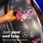 Hand pouring pink scent booster beads from a measuring cup into the drum of a washing machine with text 'Just pour and toss.'