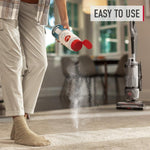 Hand pouring carpet refresher powder from a Hoover container onto carpet with an upright vacuum nearby