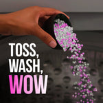 Hand pouring pink and white laundry scent beads from a measuring cup into a washing machine with text 'Toss, Wash, Wow'
