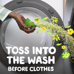 Hand pouring small yellow scent beads from a green measuring cup into a washing machine drum with floral accents.