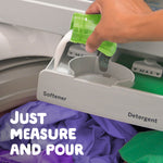 Hand pouring white liquid softener from a green cap into a washing machine dispenser labeled 'Softener', with purple laundry visible below.