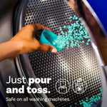 Hand pouring teal scent booster beads from a measuring scoop into a front-load washing machine drum with text ‘Just pour and toss.’