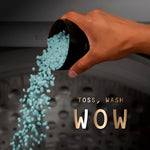 Hand pouring teal laundry scent beads from a cup into a washing machine drum with ‘Toss, Wash WOW’ text.
