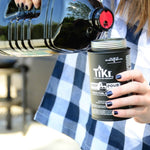 Person pouring TIKI torch fuel from a dark jug into a TIKI easy-pour canister outdoors, close-up on hands and liquid