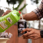 Hands pouring green TIKI BiteFighter Clean Burn torch fuel from bottle into a black TIKI pour canister outdoors
