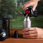 Hands pouring torch fuel from a black Mainstays bottle into a metal fuel canister on a table