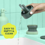Hand pouring water from the removable base of the Brite drain-and-dry stand into a sink, scrubber and lid visible