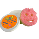 Opened Scrub Daddy PowerPaste tub with lid showing label and pink smiling Scrub Mommy sponge leaning against it