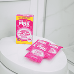 Styled product photo of The Pink Stuff box on a closed toilet lid with three pink sachets arranged beside it.