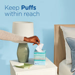 Nightstand scene with hand grabbing a tissue from a Puffs Plus Lotion cube box next to a lamp and pillow