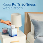 Bedside scene with a small decorative Puffs tissue box on a nightstand and a hand reaching to pull a tissue