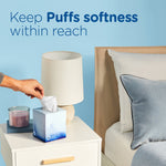 Nightstand scene with a Puffs Ultra Soft cube on a table as a hand reaches to grab a tissue beside a lamp and bed