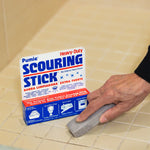 Hand using a Pumie scouring stick on tiled grout beside a standing product package with prominent branding