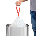 Person lifting a tied white trash bag by its red drawstrings above a stainless steel trash can.