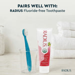 Lifestyle pairing photo showing a blue RADIUS Totz toothbrush standing in a clear glass next to a tube of RADIUS Dragon Fruit fluoride-free toothpaste on a bathroom surface.