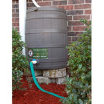 Plastic rain barrel beside a brick wall with a green garden hose attached and running across red mulch
