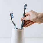 Two Reach toothbrushes placed in a ceramic holder with a hand reaching for one, styled as a simple lifestyle product shot.