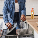 Person recycling a plastic bottle at a kitchen counter with a Febreze Luxe Tropical Blend can visible on the countertop