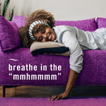 Woman smiling and resting on a purple couch with overlaid text 'breathe in the "mmhmmm"' promoting fabric freshness.