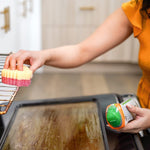 Person removing an oven rack holding a yellow-and-pink Scrub Daddy sponge and a Cif bottle in the other hand