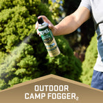 Hand holding and spraying a Repel Camp Fogger aerosol can outdoors with visible spray mist and green foliage background