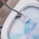 Grey toilet brush scrubbing underside rim of a toilet bowl with blue soapy water visible