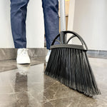 Low-angle lifestyle shot of a Reynera broom head sweeping a glossy dark tile floor with a person's legs visible.