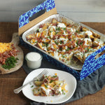 Styled tabletop photo of a loaded baked dish in a Reynolds aluminum pan placed inside a blue patterned carrier with a plated serving