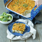 Lifestyle shot of a baked casserole in a Reynolds disposable aluminum pan inside a decorative carrier and a served square on a plate