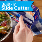 Hand using a built-in slide cutter on a Reynolds Kitchens plastic wrap box with a salad bowl beside it and 'Built-In Slide Cutter' banner