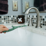 Hand holding a green Dr. Brown's toddler toothbrush under a running bathroom sink faucet to rinse bristles.
