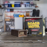 Roach Motel box and an opened cardboard trap displayed on a metal shelf in a garage setting.