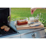 Outdoor lifestyle scene showing a Rubbermaid Brilliance rectangular container on a cutting board next to a grill, being seasoned by a hand.
