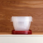 Clear square Rubbermaid containers stacked without lids with a pile of red lids beneath on a wooden shelf